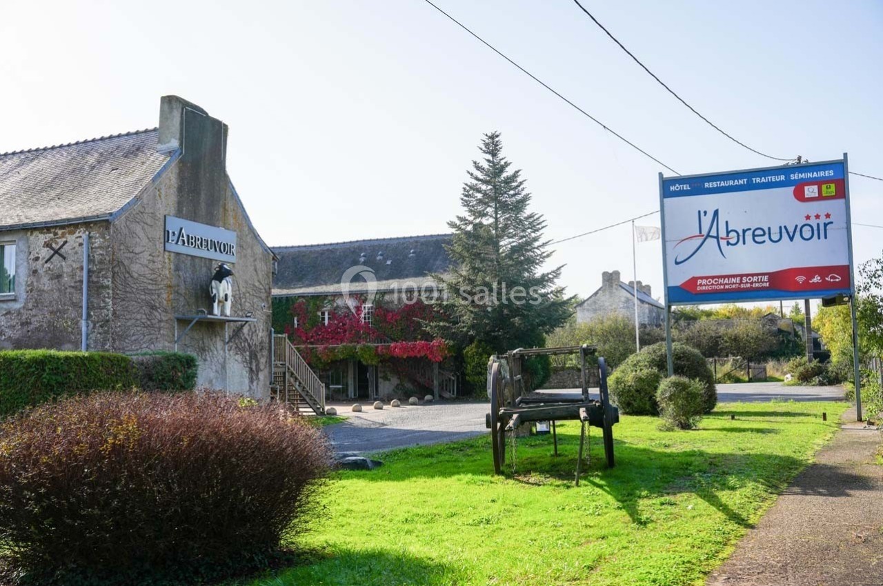 Façade d'un bâtiment en pierre avec enseigne ’L'Abreuvoir’, entouré de verdure et d'un panneau publicitaire.