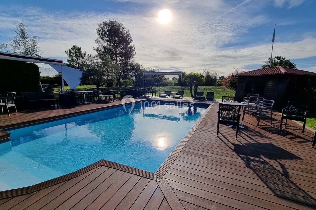 Piscine extérieure entourée d'une terrasse en bois, avec mobilier de jardin et végétation sous un ciel ensoleillé.