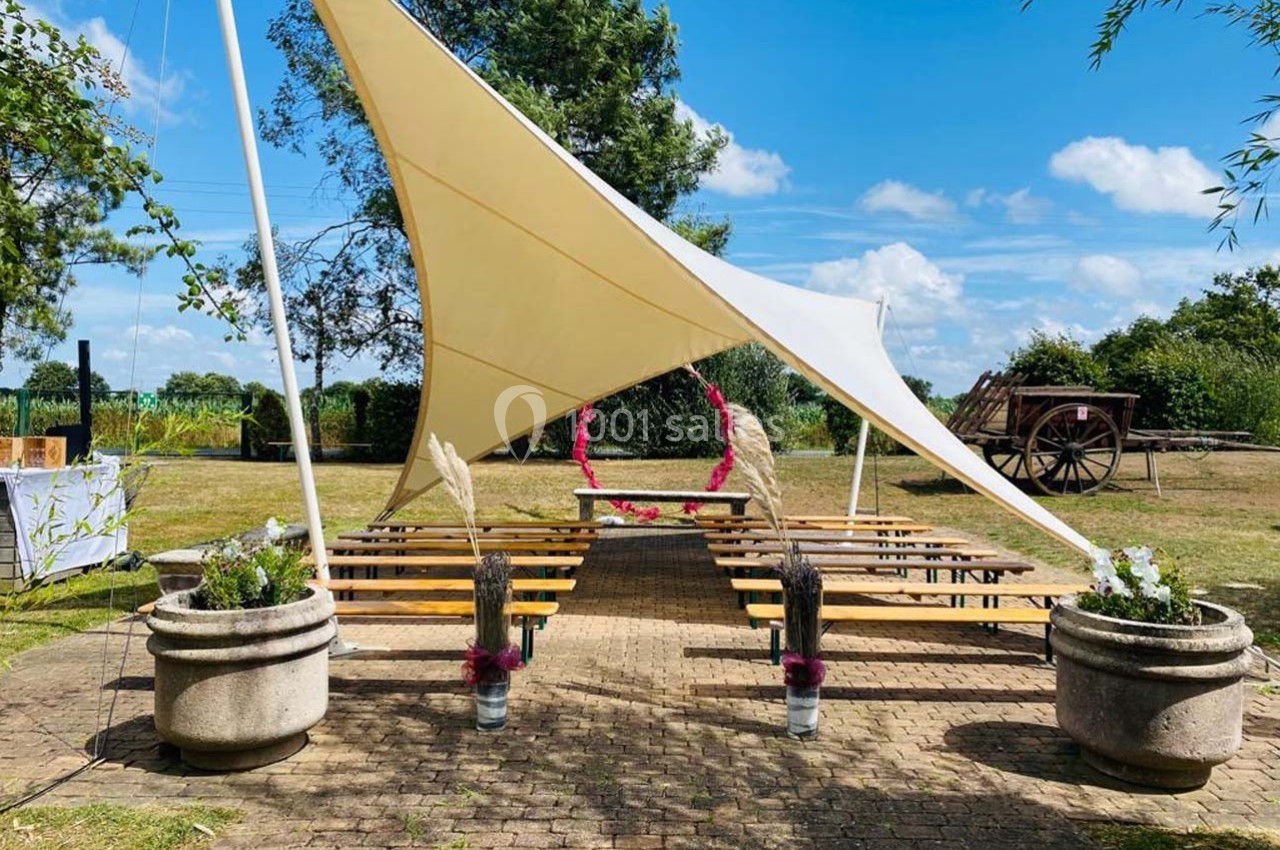Espace extérieur aménagé avec une tente blanche, des bancs en bois et des décorations florales, dans un cadre champêtre.
