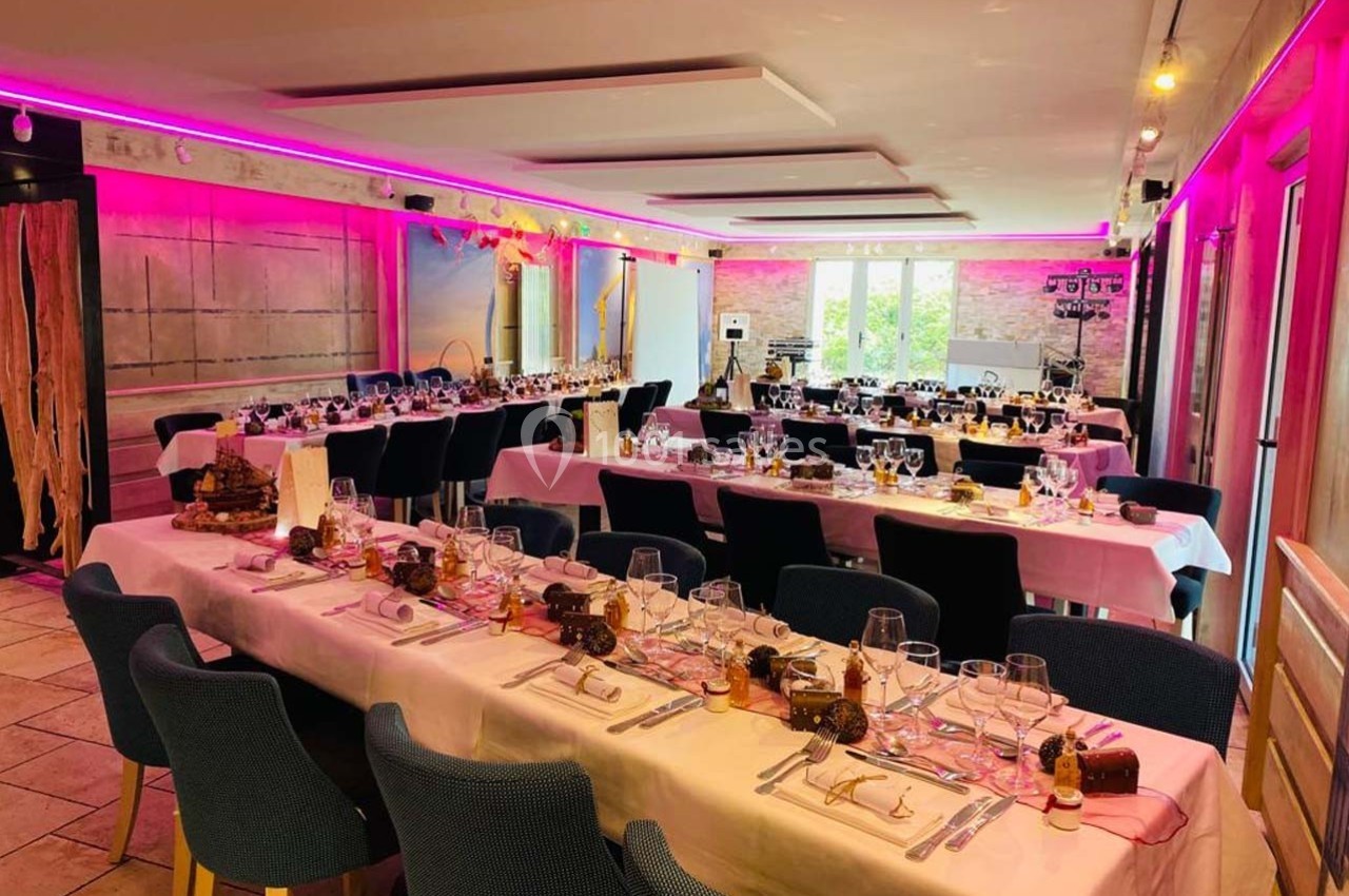 Salle décorée pour un repas festif, avec tables dressées, nappes blanches, chaises bleues et éclairage rose.