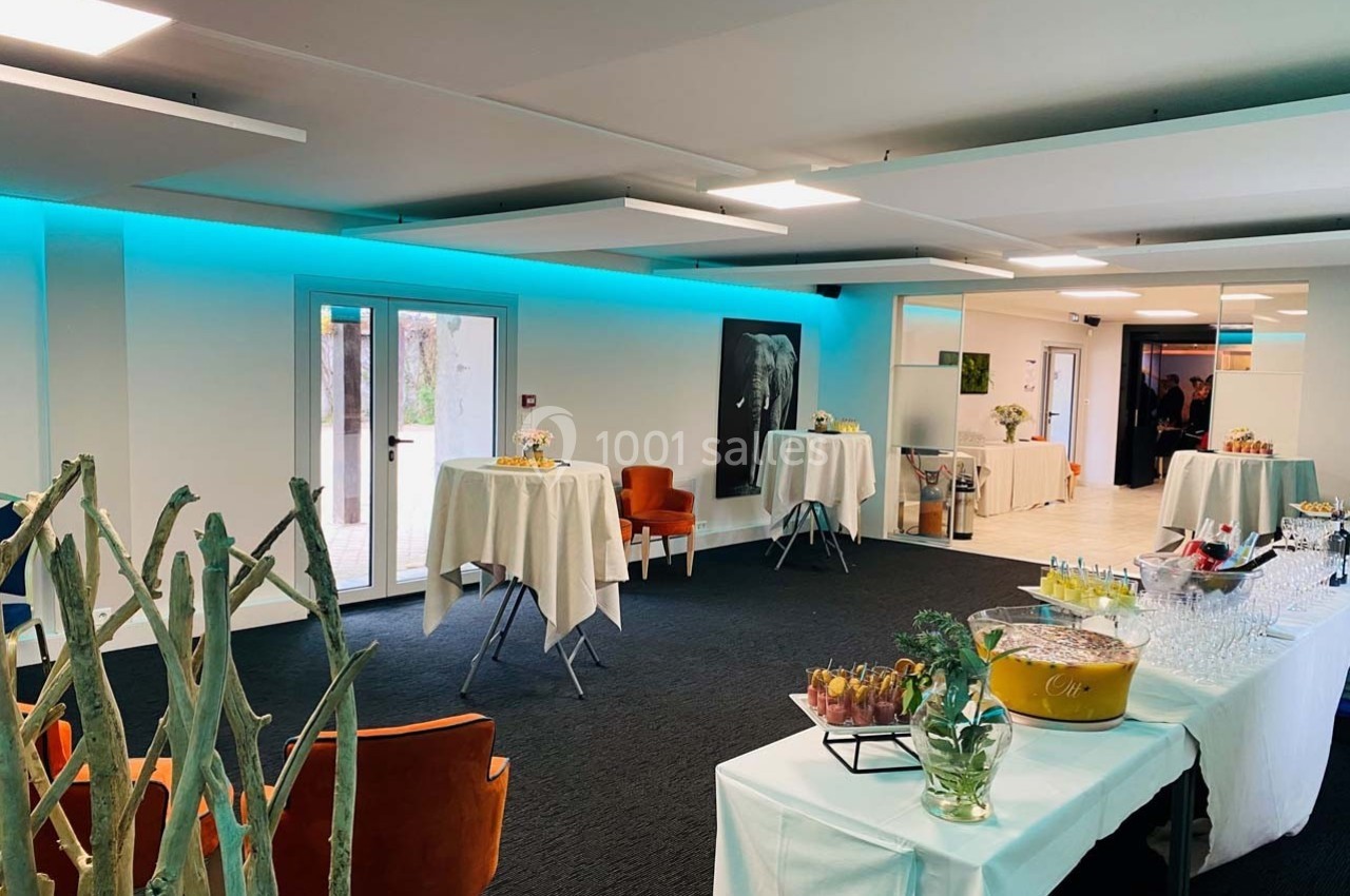 Salle lumineuse aménagée pour un événement avec tables hautes, buffet, et décoration moderne.