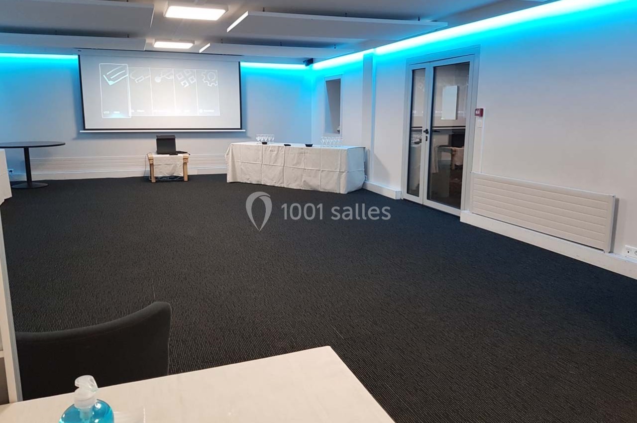 Salle de réunion vide avec un projecteur allumé, des tables couvertes de nappes blanches et des chaises disposées autour.