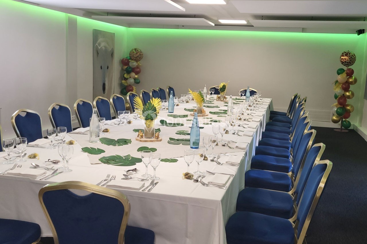 Salle de réception avec une grande table dressée, décorée de feuilles tropicales, ananas et ballons colorés en arrière-plan.