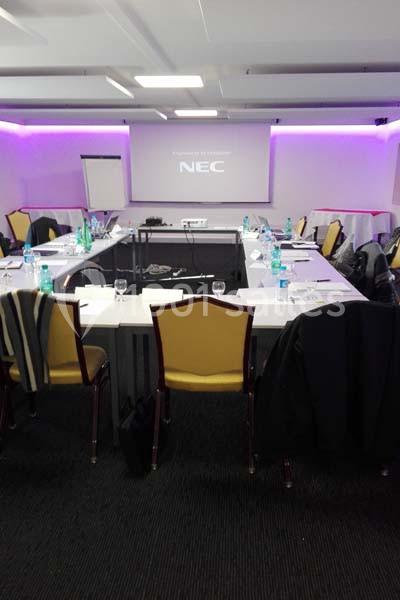 Salle de réunion avec tables disposées en U, chaises, écran de projection allumé et bouteilles d'eau sur les tables.