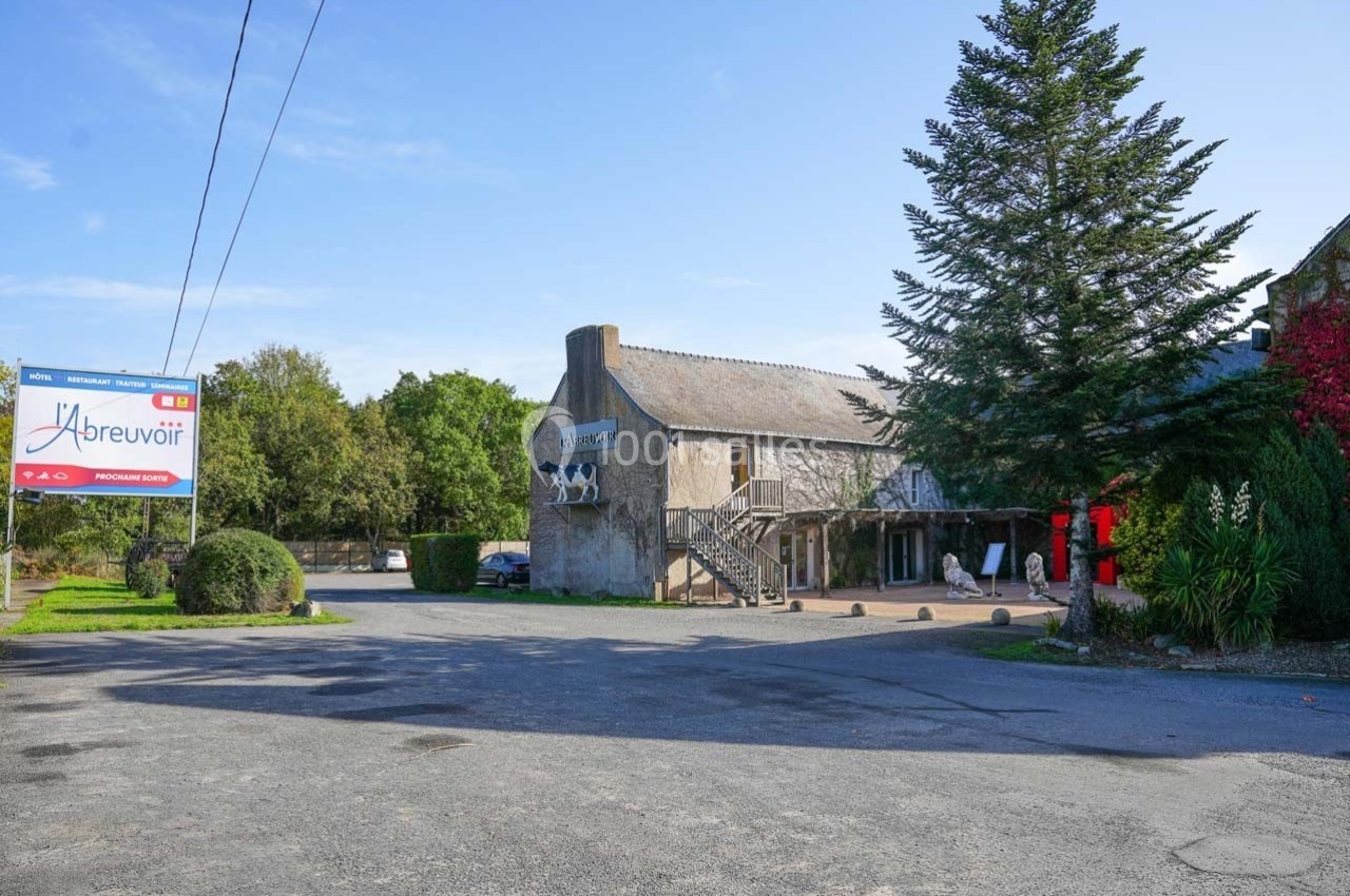 Façade d'un restaurant en pierre avec enseigne ’L'Abreuvoir’, entouré de végétation et d'un parking dégagé.