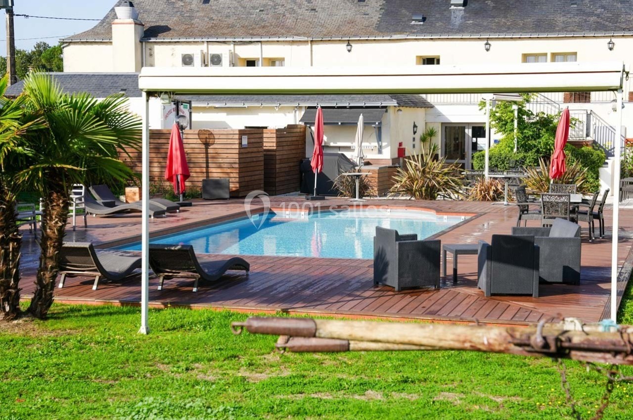 Piscine extérieure entourée de chaises longues, parasols rouges et terrasse en bois, avec bâtiment en arrière-plan.