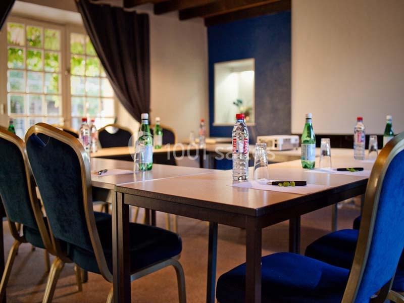 Salle de réunion aménagée avec des tables, des chaises, des bouteilles d'eau et des blocs-notes, éclairée par une fenêtre.