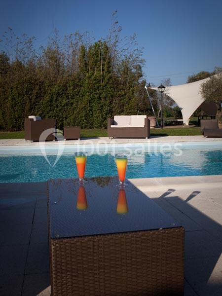 Deux verres de cocktail colorés posés sur une table en rotin près d'une piscine, avec des fauteuils et un jardin en arrière…