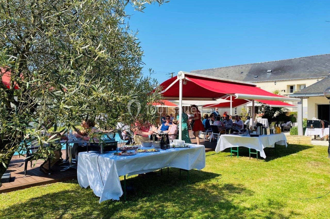 Des tables dressées sous des parasols rouges dans un jardin ensoleillé, avec des convives assis et debout.