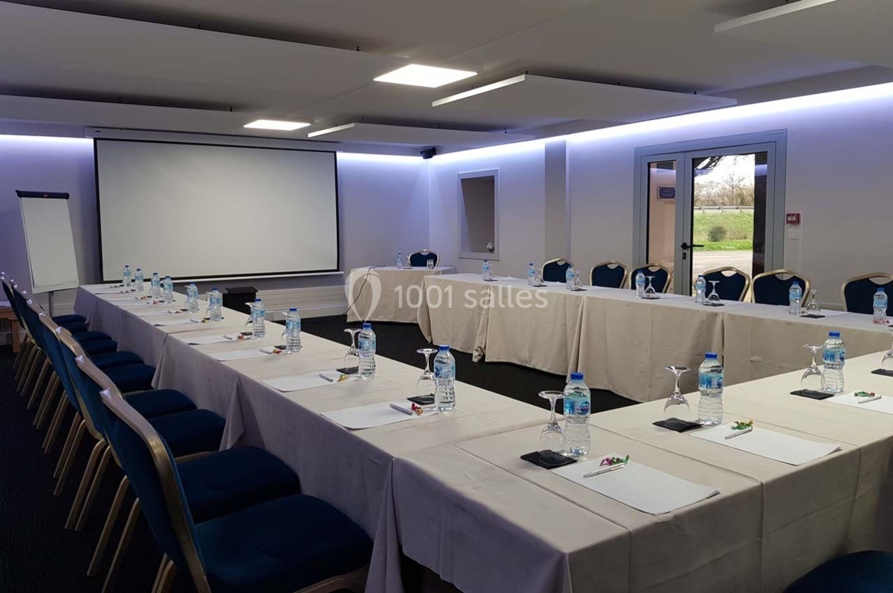 Salle de réunion avec tables disposées en U, chaises alignées, écran de projection, paperboard et bouteilles d'eau.