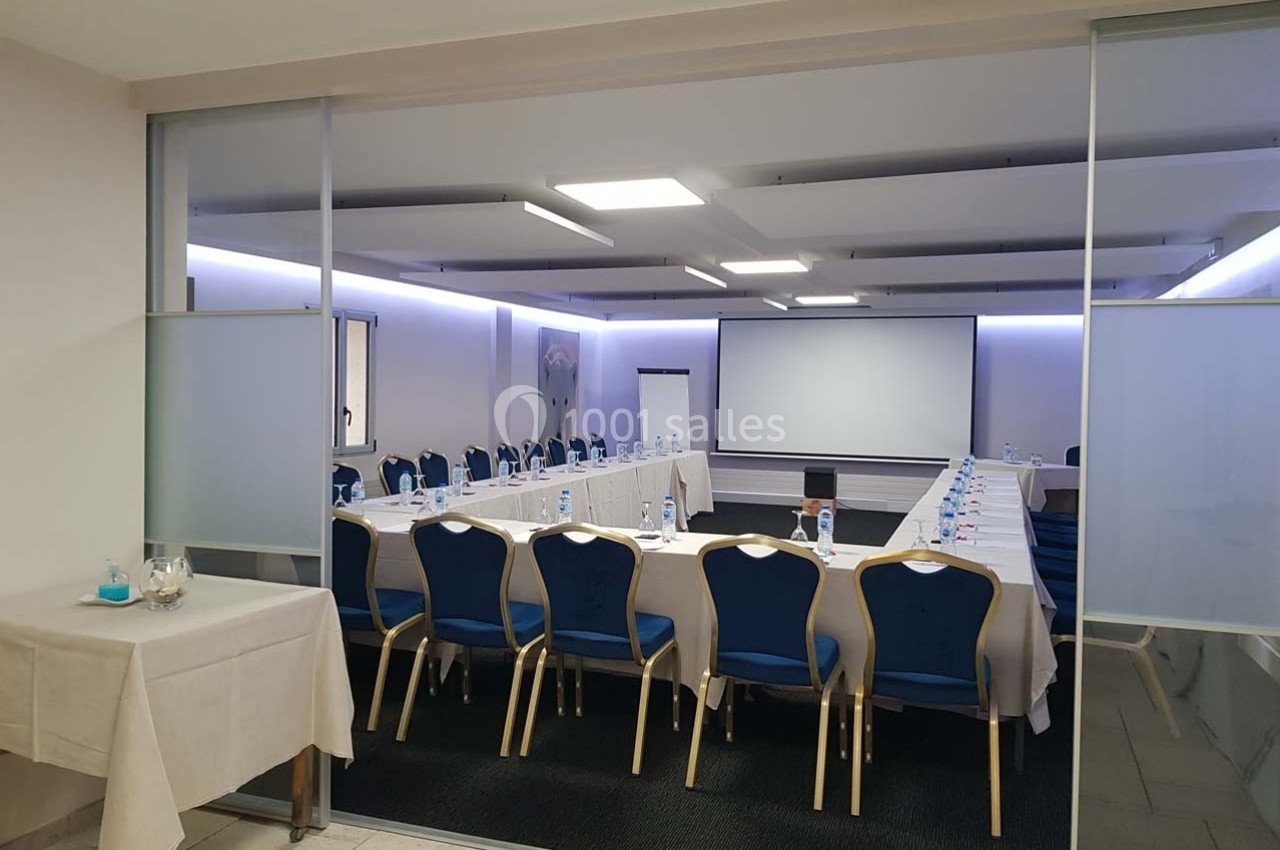 Salle de réunion équipée de tables en U, chaises bleues, écran de projection et éclairage moderne.