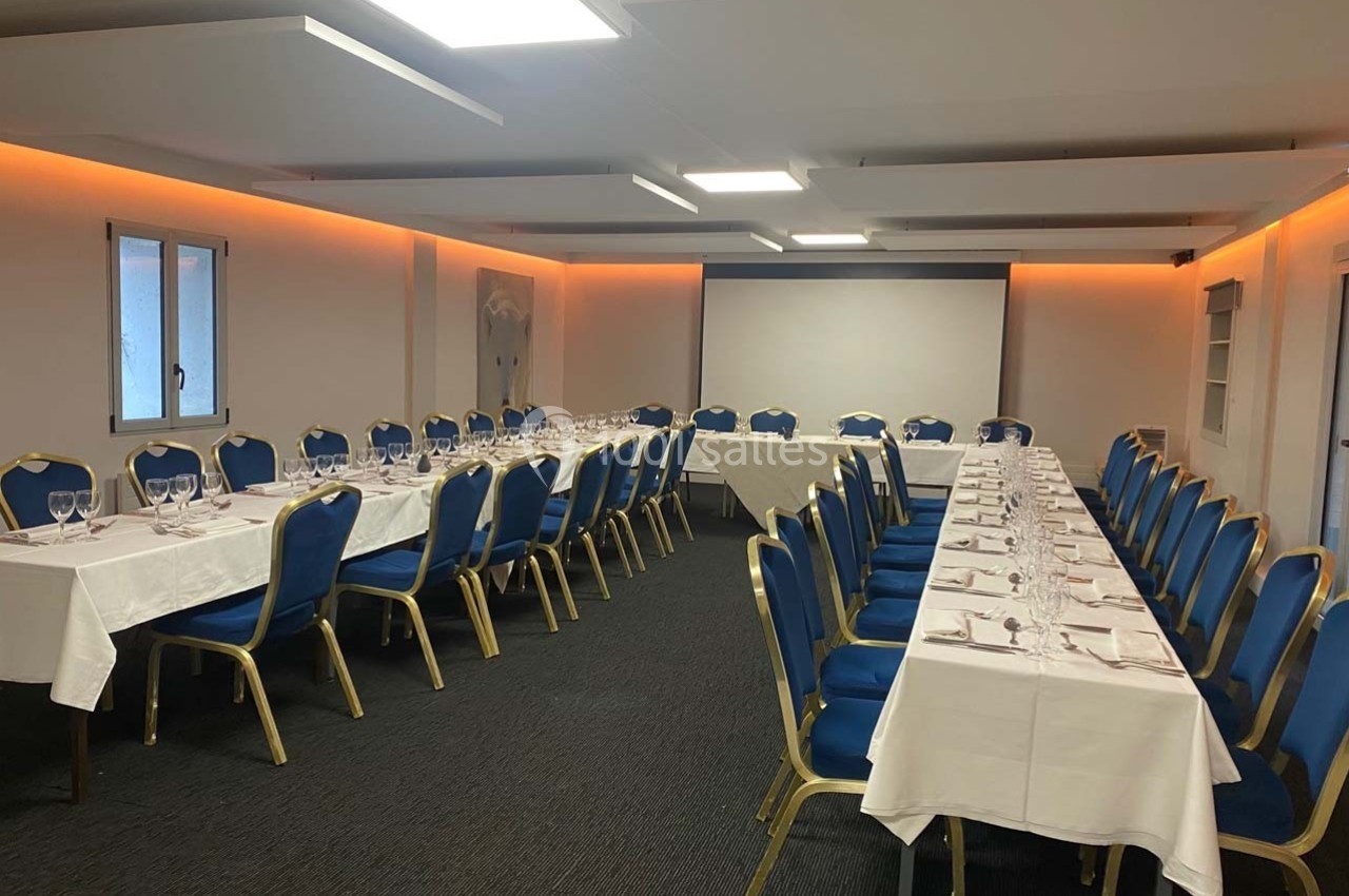 Salle de réunion aménagée en U avec des tables blanches, chaises bleues et un écran de projection en fond.
