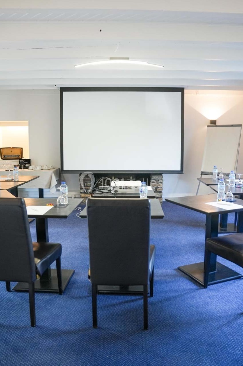 Salle de réunion équipée de tables, chaises, écran de projection, paperboard et bouteilles d'eau sur moquette bleue.