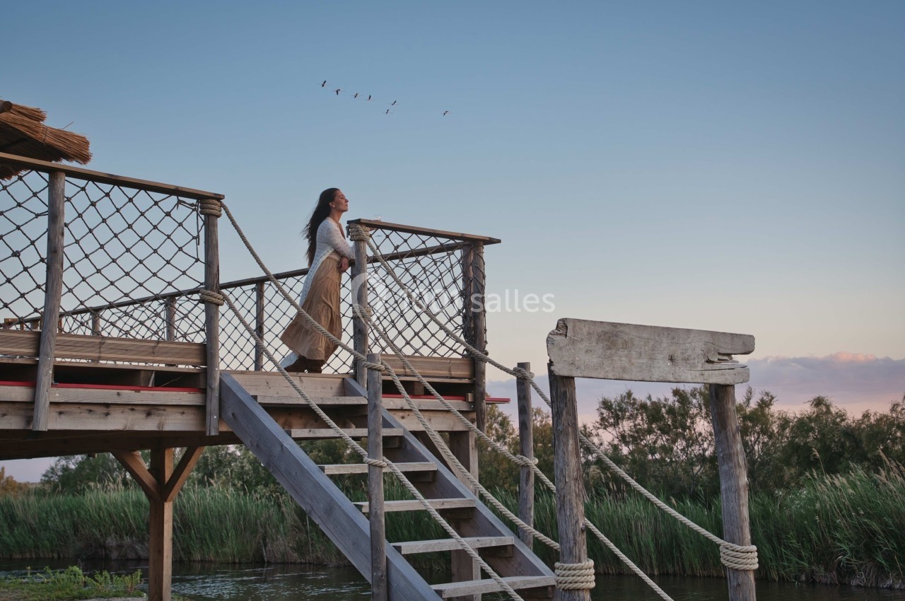 Une femme debout sur une plateforme en bois, regardant l'horizon au-dessus d'un paysage naturel avec des roseaux.