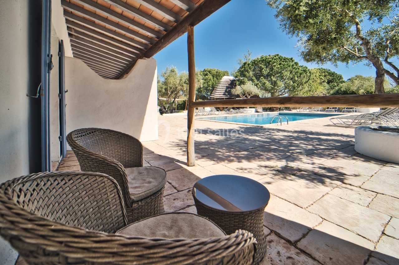 Terrasse avec fauteuils en rotin, table basse, vue sur une piscine entourée de végétation et ciel dégagé.