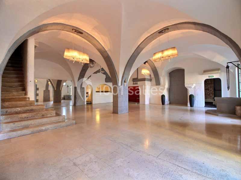 Intérieur spacieux avec arches en pierre, escalier à gauche, sol en marbre et luminaires suspendus.