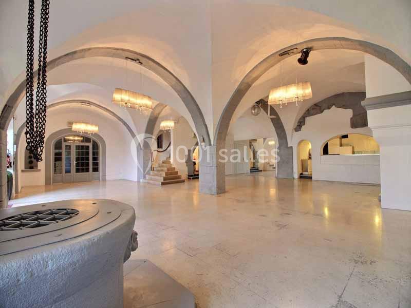 Salle voûtée lumineuse avec des arches en pierre, un puits au premier plan et des lustres suspendus.