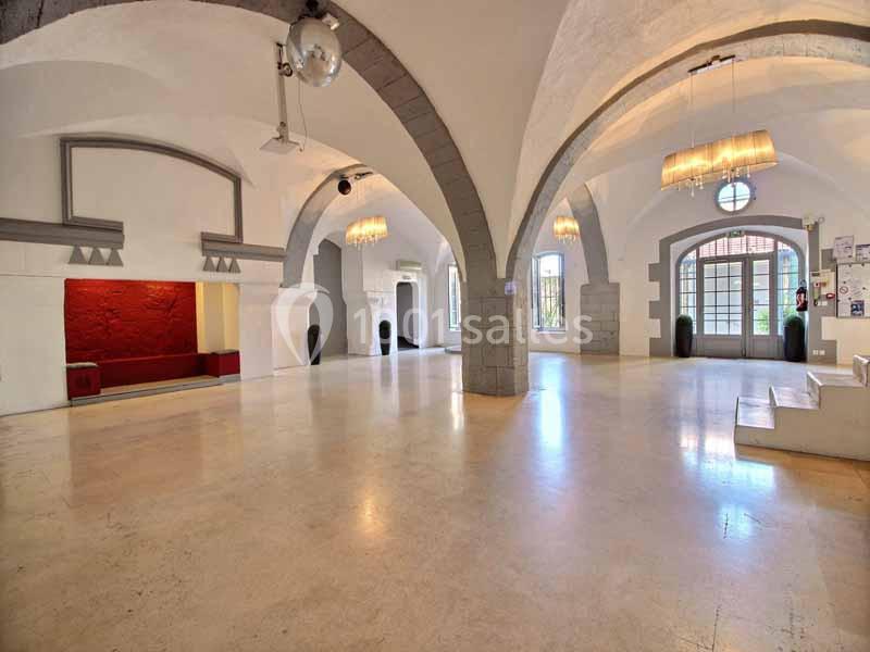Salle lumineuse avec arches en pierre, sol clair, murs blancs, espace rouge en alcôve et portes vitrées au fond.