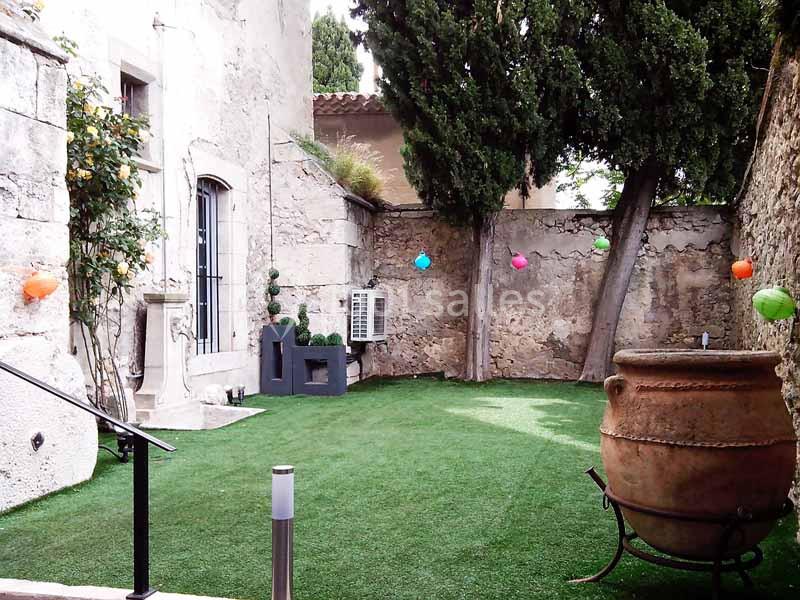 Cour intérieure avec pelouse artificielle, murs en pierre, arbres, pots décoratifs et guirlandes de lampions colorés.