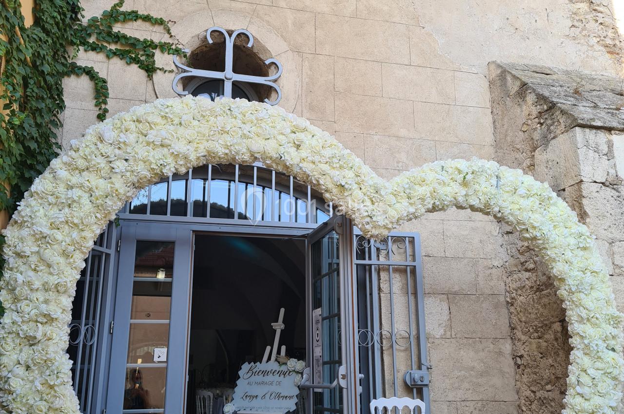 Entrée décorée d'une arche en forme de cœur composée de fleurs blanches, devant un bâtiment en pierre.