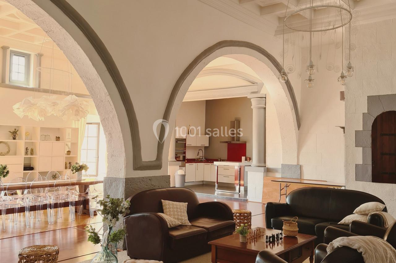 Salon lumineux avec canapés en cuir, table basse en bois, arches en pierre et vue sur une cuisine ouverte.