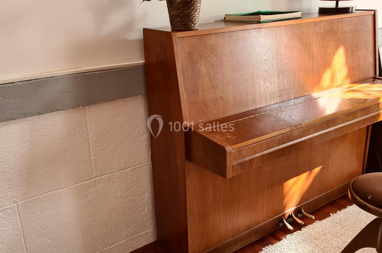 Un piano droit en bois avec une plante posée dessus, un échiquier au sol et un tapis clair dans une pièce lumineuse.