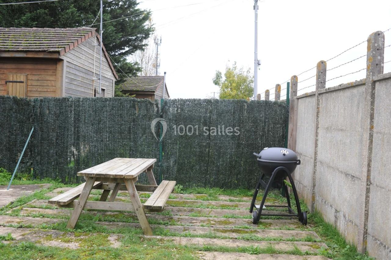 Table de pique-nique en bois et barbecue sur une terrasse extérieure entourée de clôtures et de végétation.