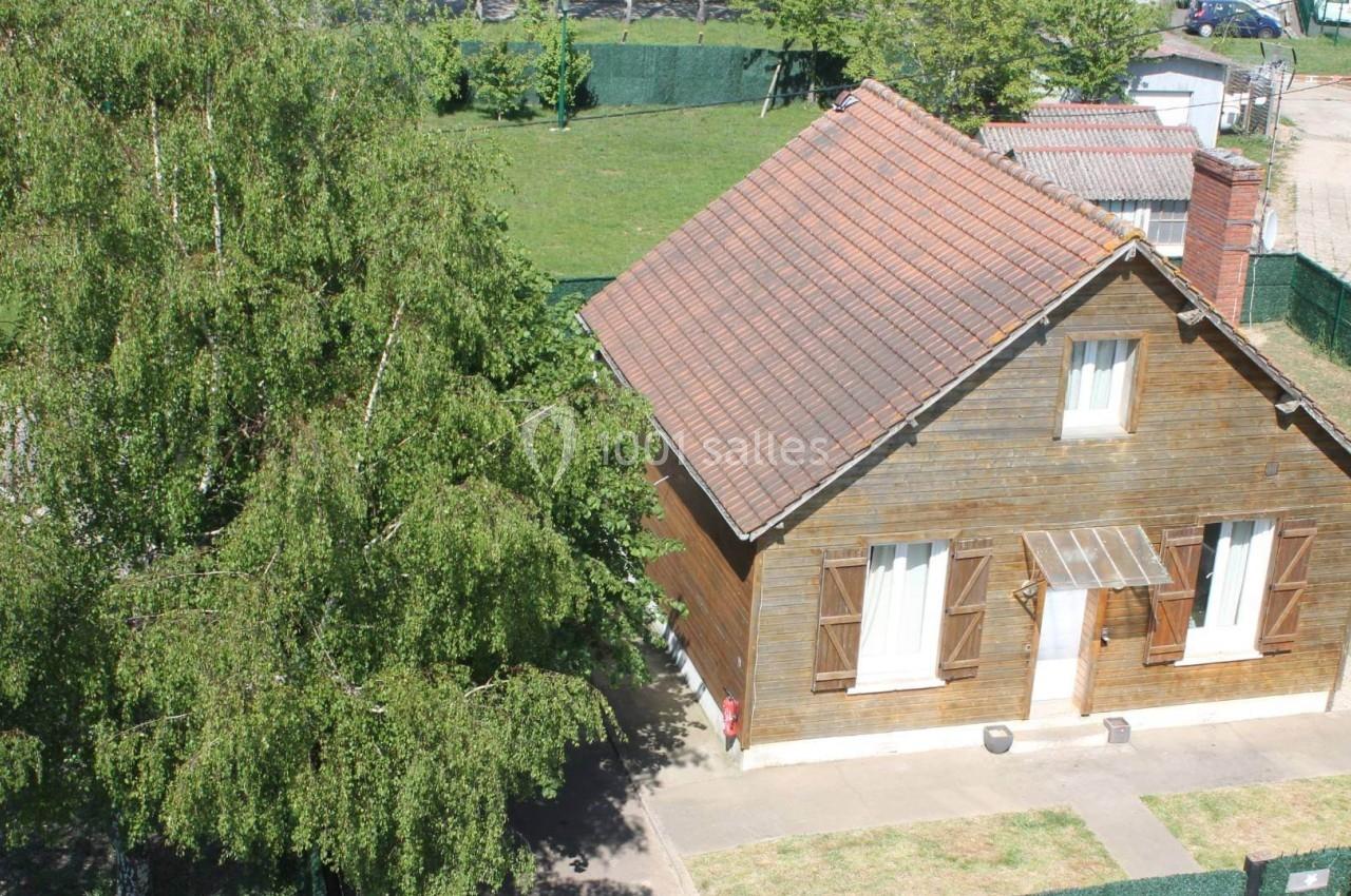Maison en bois avec toit en tuiles rouges, entourée d'un jardin arboré et clôturé, vue en journée ensoleillée.