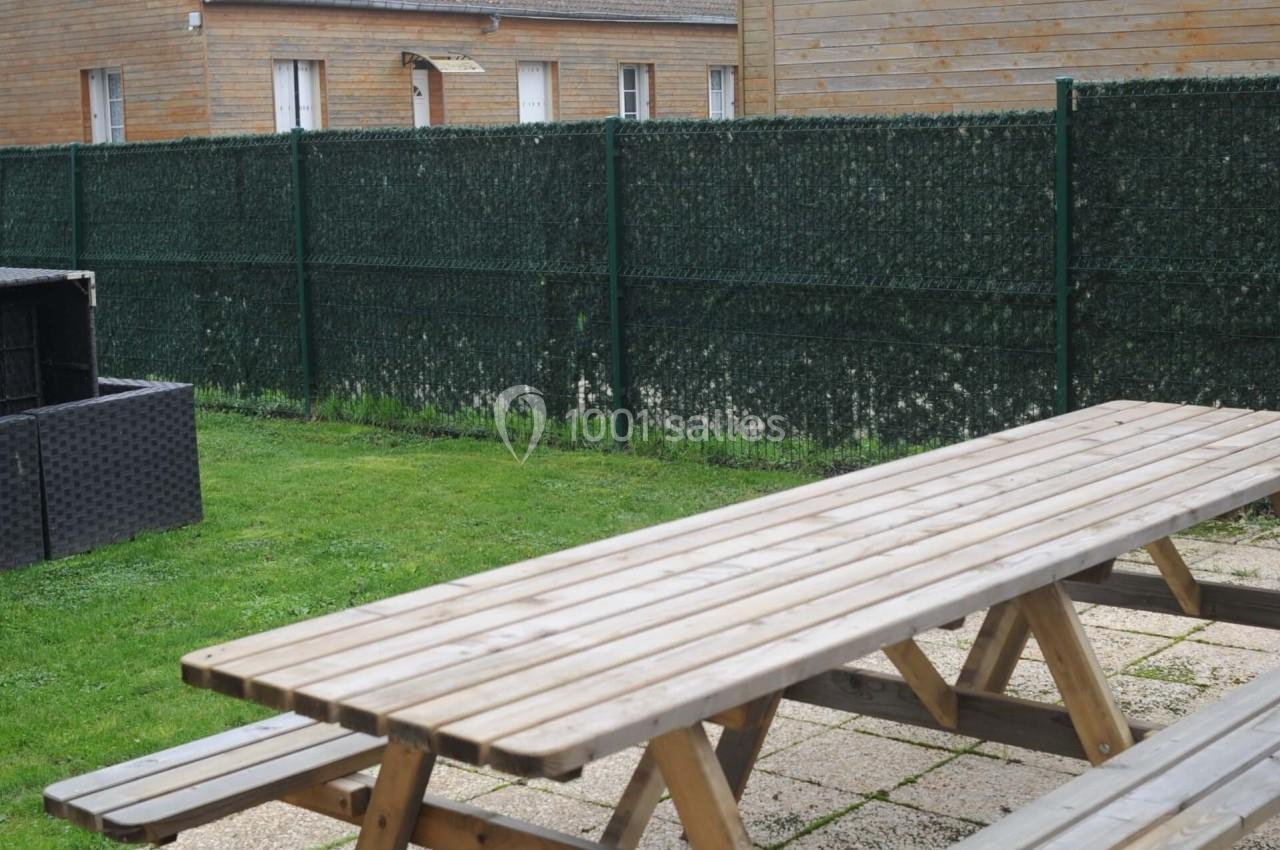 Table de pique-nique en bois sur une terrasse pavée, entourée de pelouse et clôturée par un grillage vert.