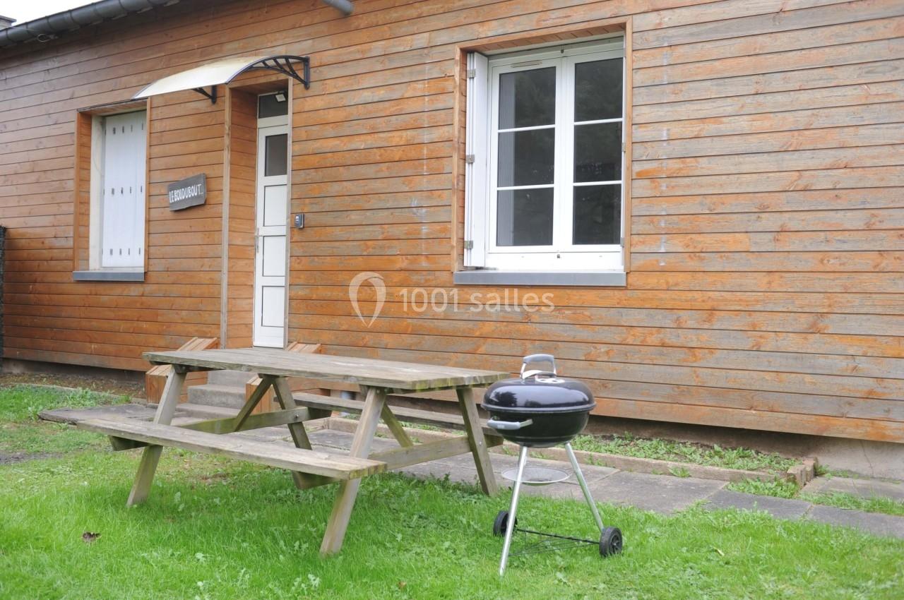 Table de pique-nique en bois et barbecue sur pelouse devant une maison en bois avec porte et fenêtre blanches.