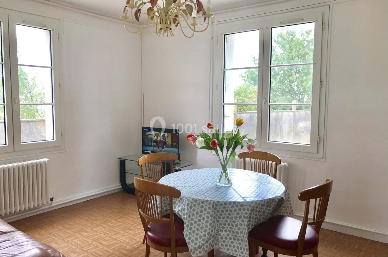 Salle à manger lumineuse avec table ronde, chaises en bois, vase de fleurs et fenêtres donnant sur un extérieur verdoyant.