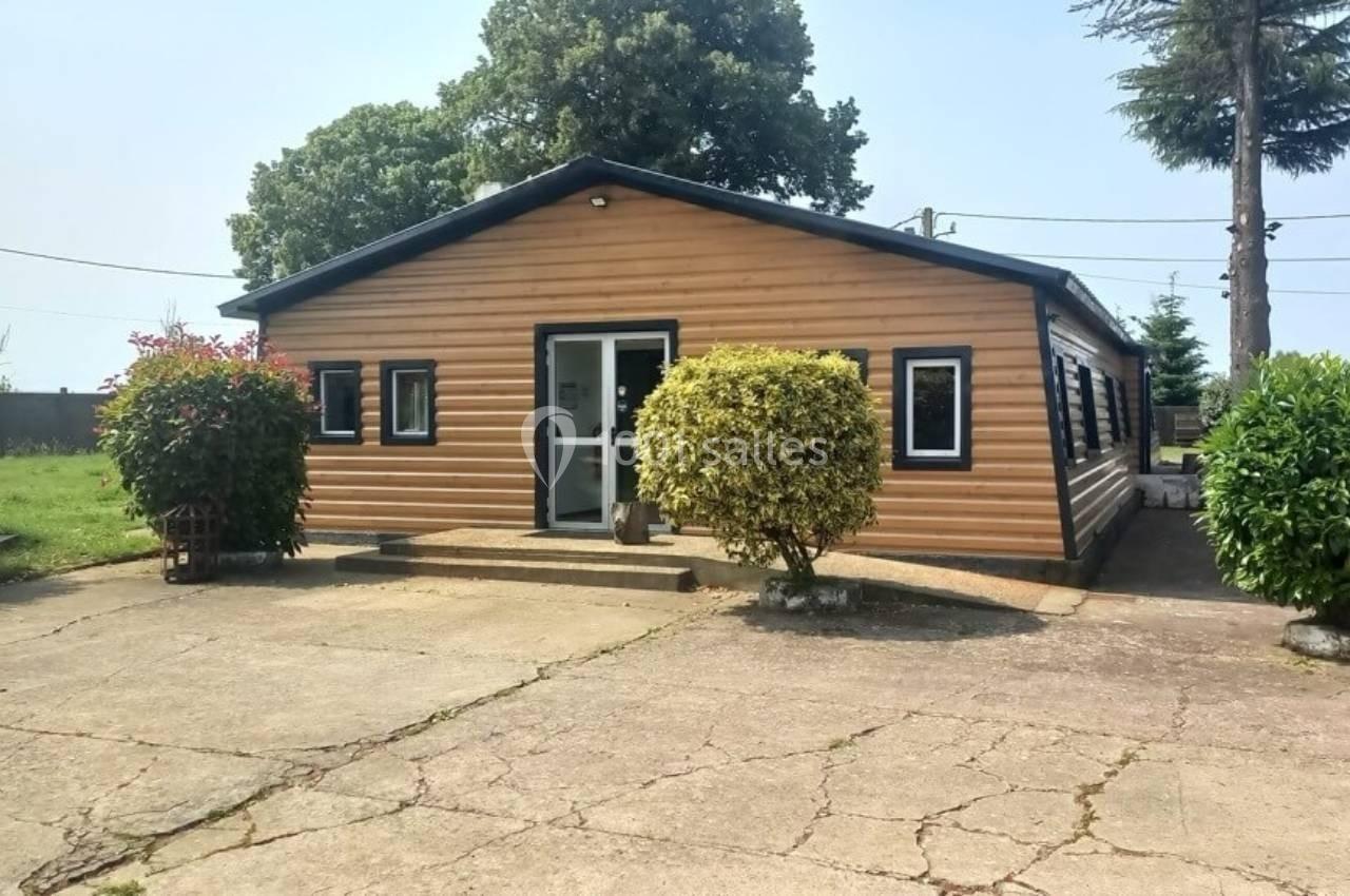 Bâtiment en bois avec plusieurs fenêtres, entouré de végétation et situé sur une surface bétonnée.