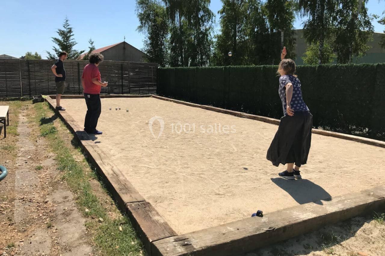 Trois personnes jouent à la pétanque sur un terrain ensoleillé entouré de haies et de clôtures.