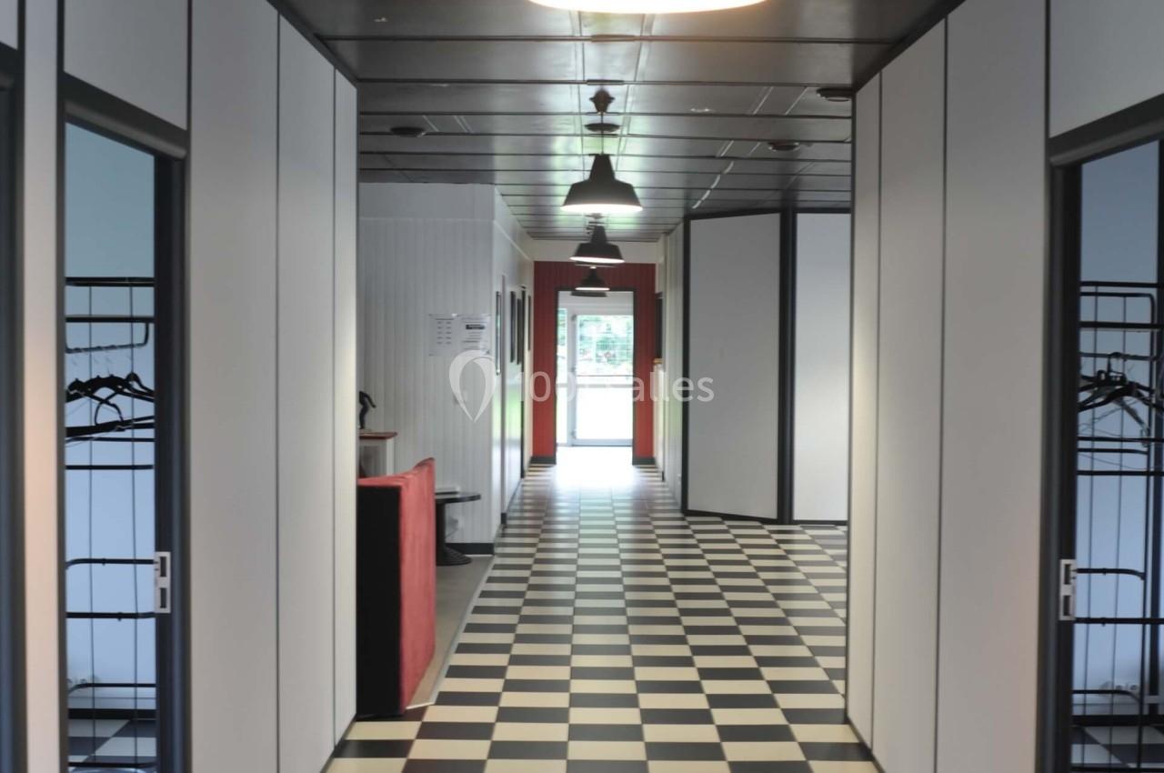 Couloir avec sol en damier noir et blanc, murs blancs, portes grises et éclairage suspendu.