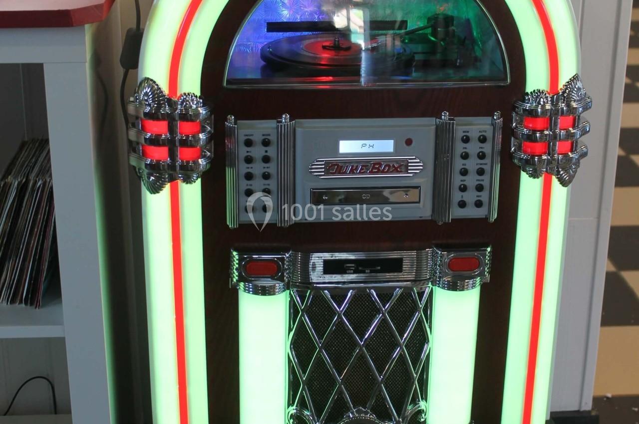 Jukebox rétro lumineux avec façade incurvée, posé sur un sol en moquette devant un mur blanc.