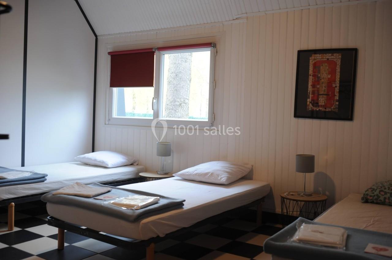 Chambre lumineuse avec trois lits simples, draps pliés, lampes de chevet et fenêtre avec store rouge.