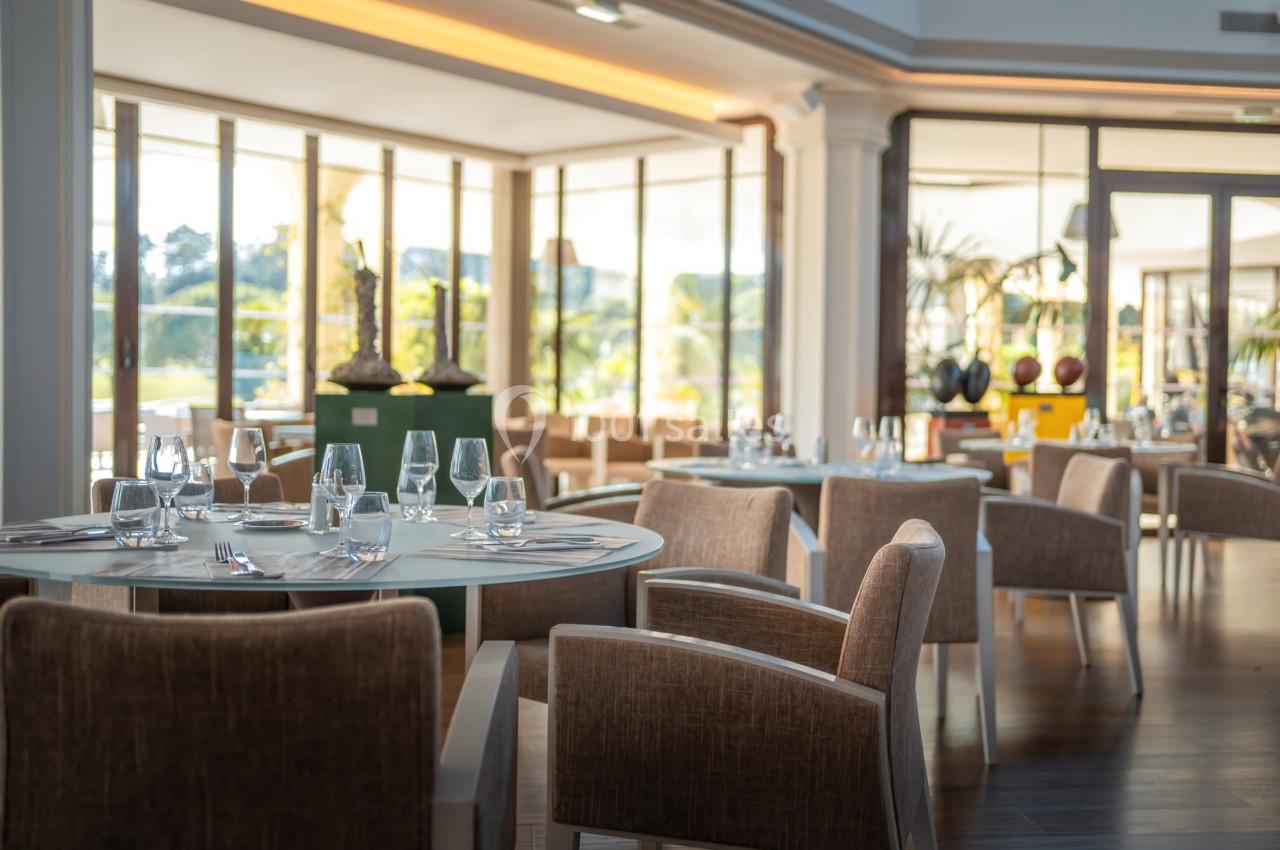 Salle de restaurant lumineuse avec tables dressées, chaises en tissu beige et grandes baies vitrées donnant sur l'extérieur.