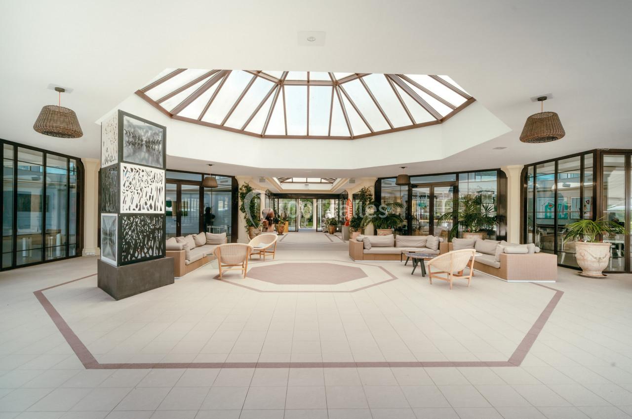 Hall spacieux avec verrière centrale, mobilier en rotin, plantes décoratives et larges baies vitrées.