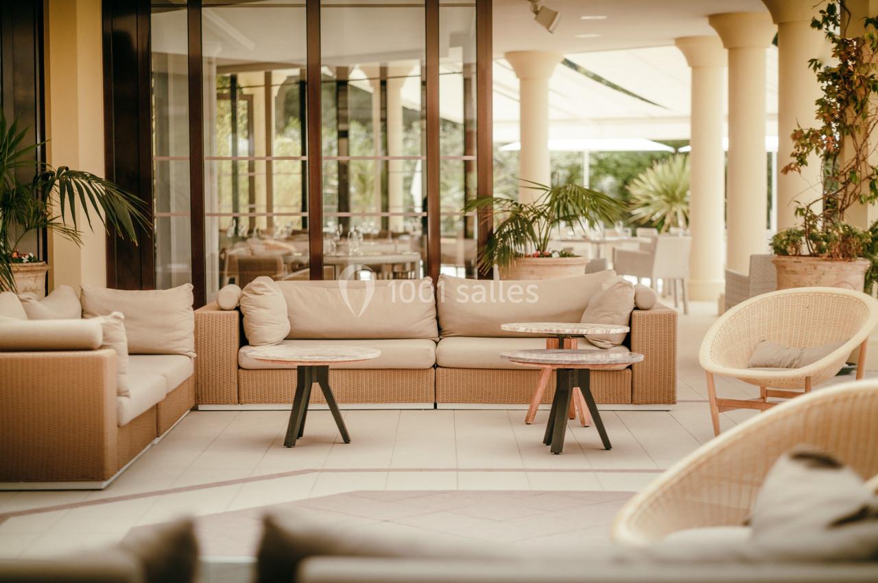 Salon extérieur avec canapés en rotin, tables basses et plantes, situé sous une pergola lumineuse.