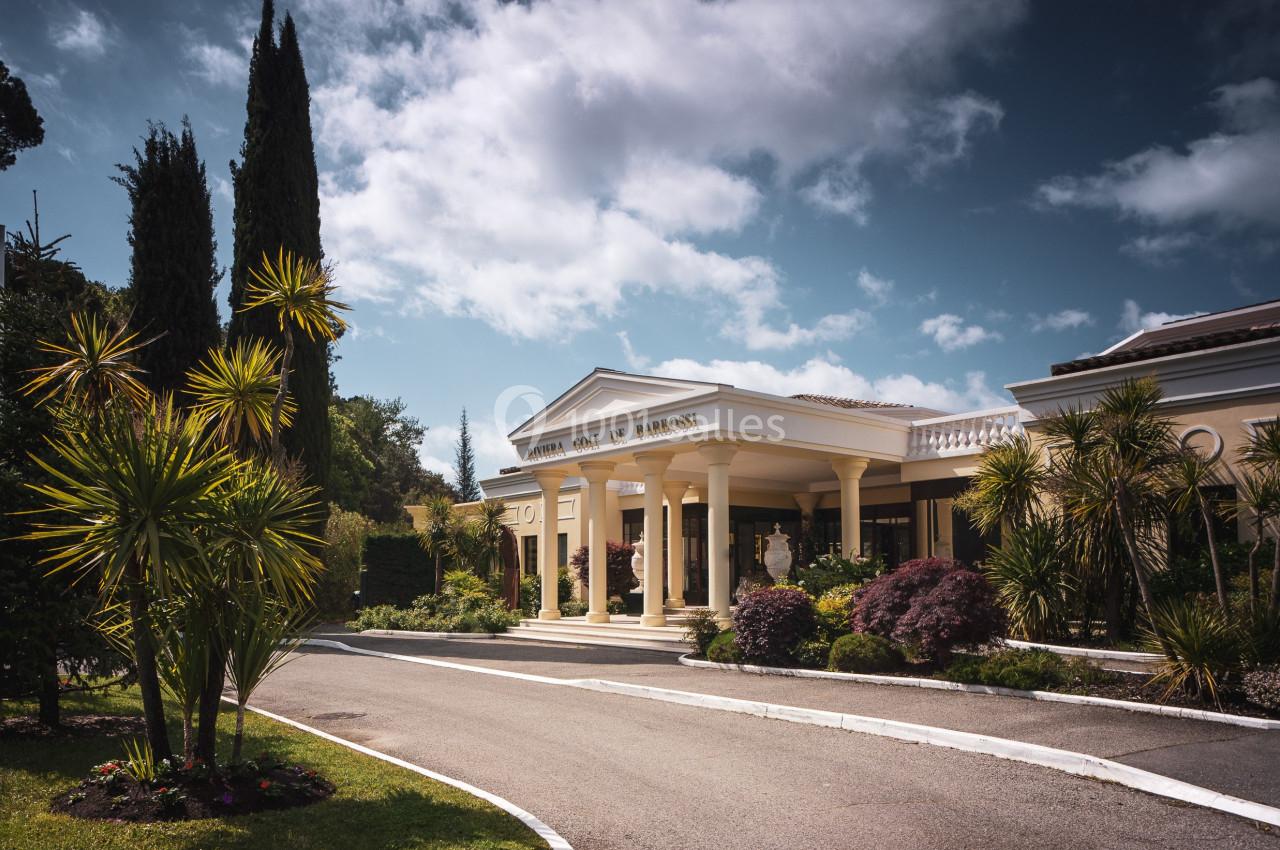 Entrée d'un bâtiment élégant entouré de végétation, avec une allée bordée de plantes et un ciel partiellement nuageux.