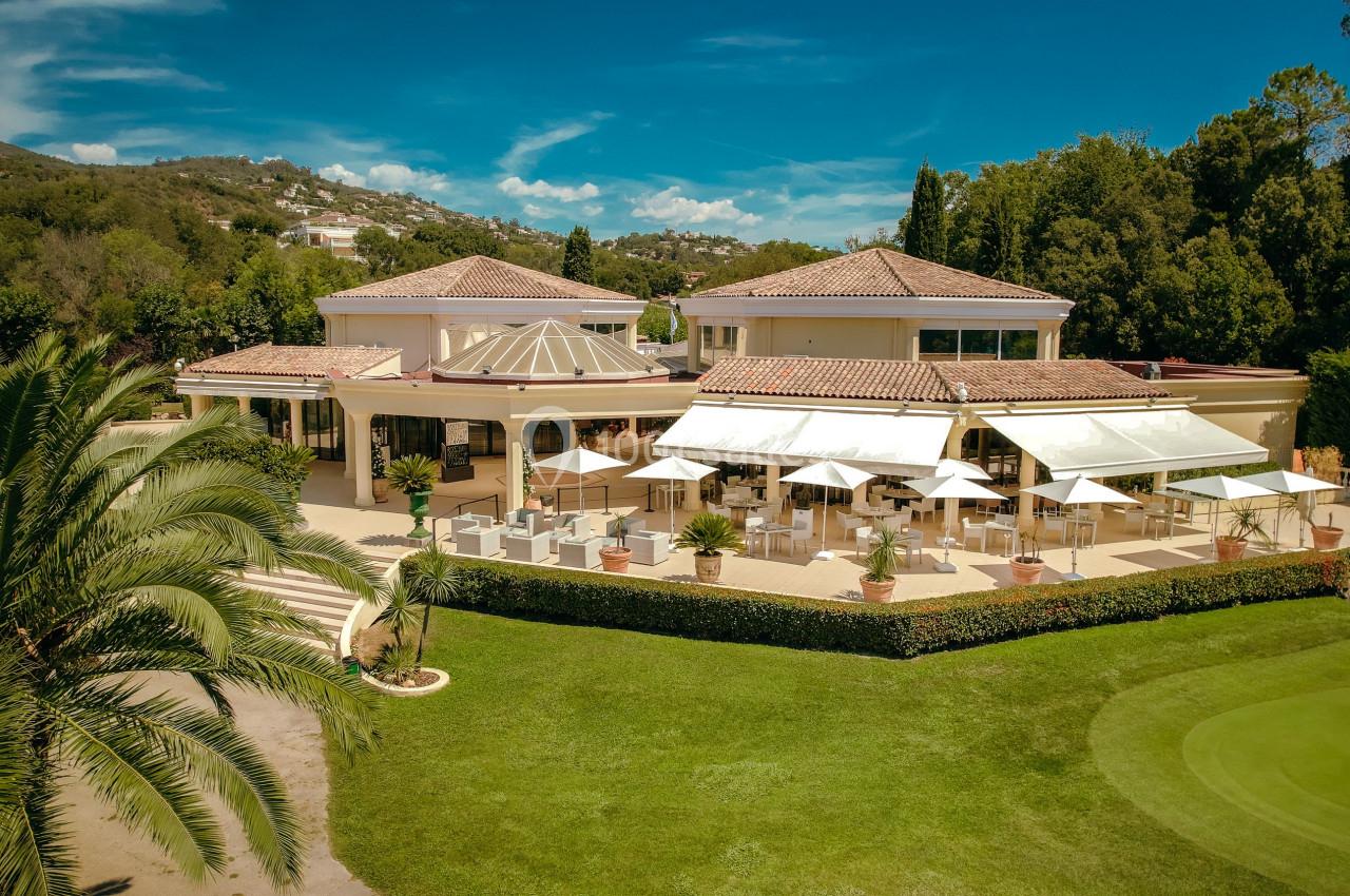 Vue aérienne d'un bâtiment lumineux entouré de verdure, avec une terrasse aménagée et des parasols blancs.