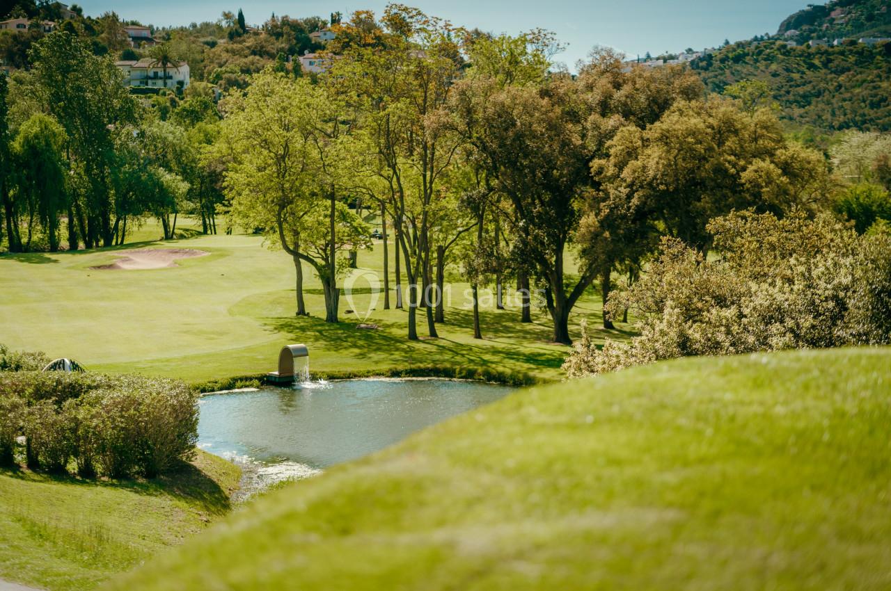 Vue d'un terrain de golf verdoyant avec un étang, des arbres et des collines en arrière-plan.