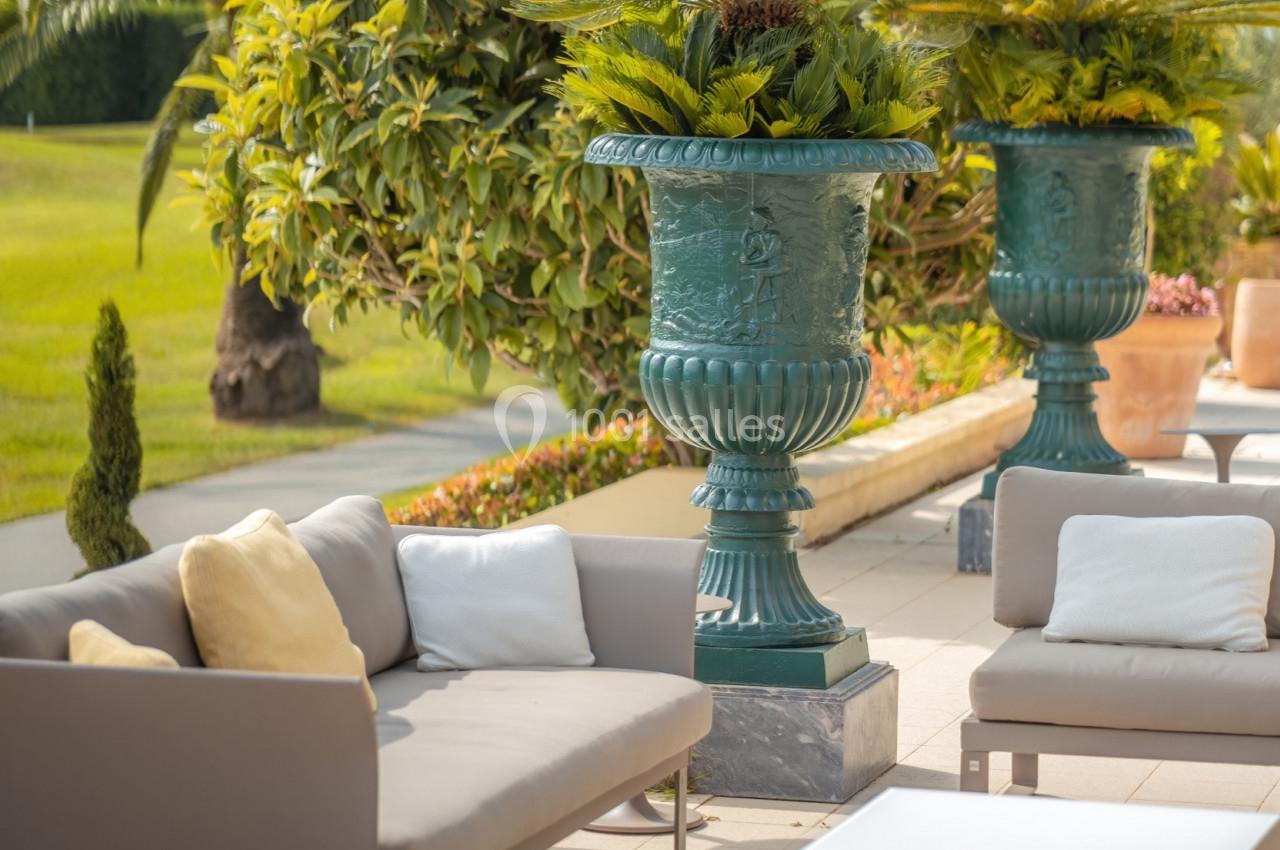 Mobilier de jardin moderne avec canapés gris et tables basses, entouré de plantes et de grands vases décoratifs.