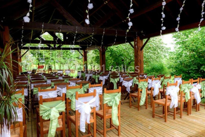 Chaises en bois décorées de rubans verts et blancs alignées sous une pergola en bois, entourée de verdure.
