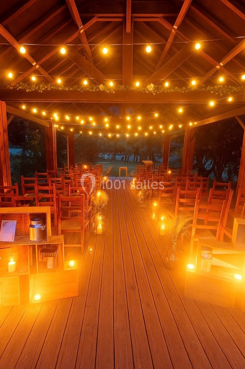 Salle en bois décorée pour un événement, avec des rangées de chaises, des guirlandes lumineuses et des bougies au sol.
