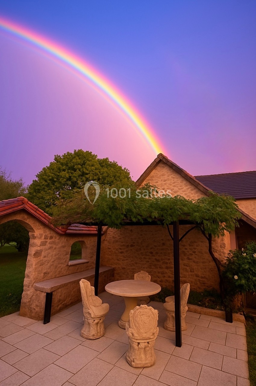 Terrasse en pierre avec table et chaises sculptées, pergola végétalisée, arc-en-ciel et ciel violet en arrière-plan.