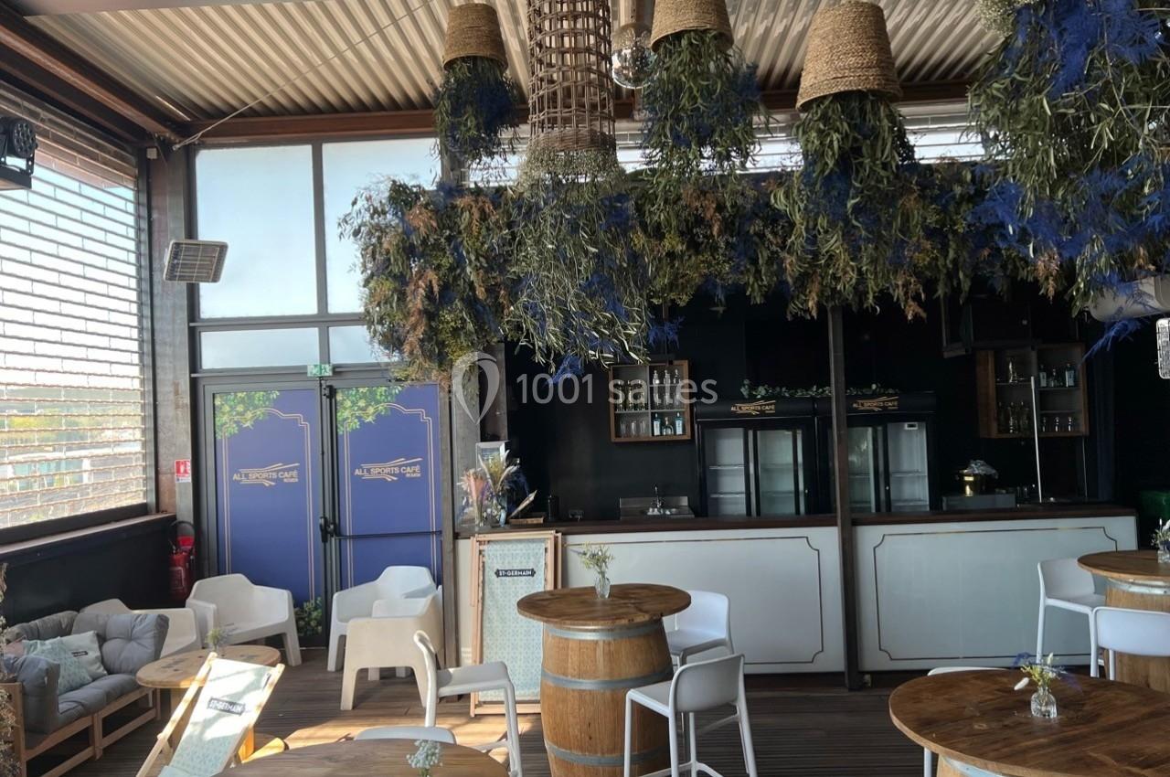 Espace intérieur lumineux avec tables en bois, chaises blanches et décoration suspendue composée de plantes séchées.