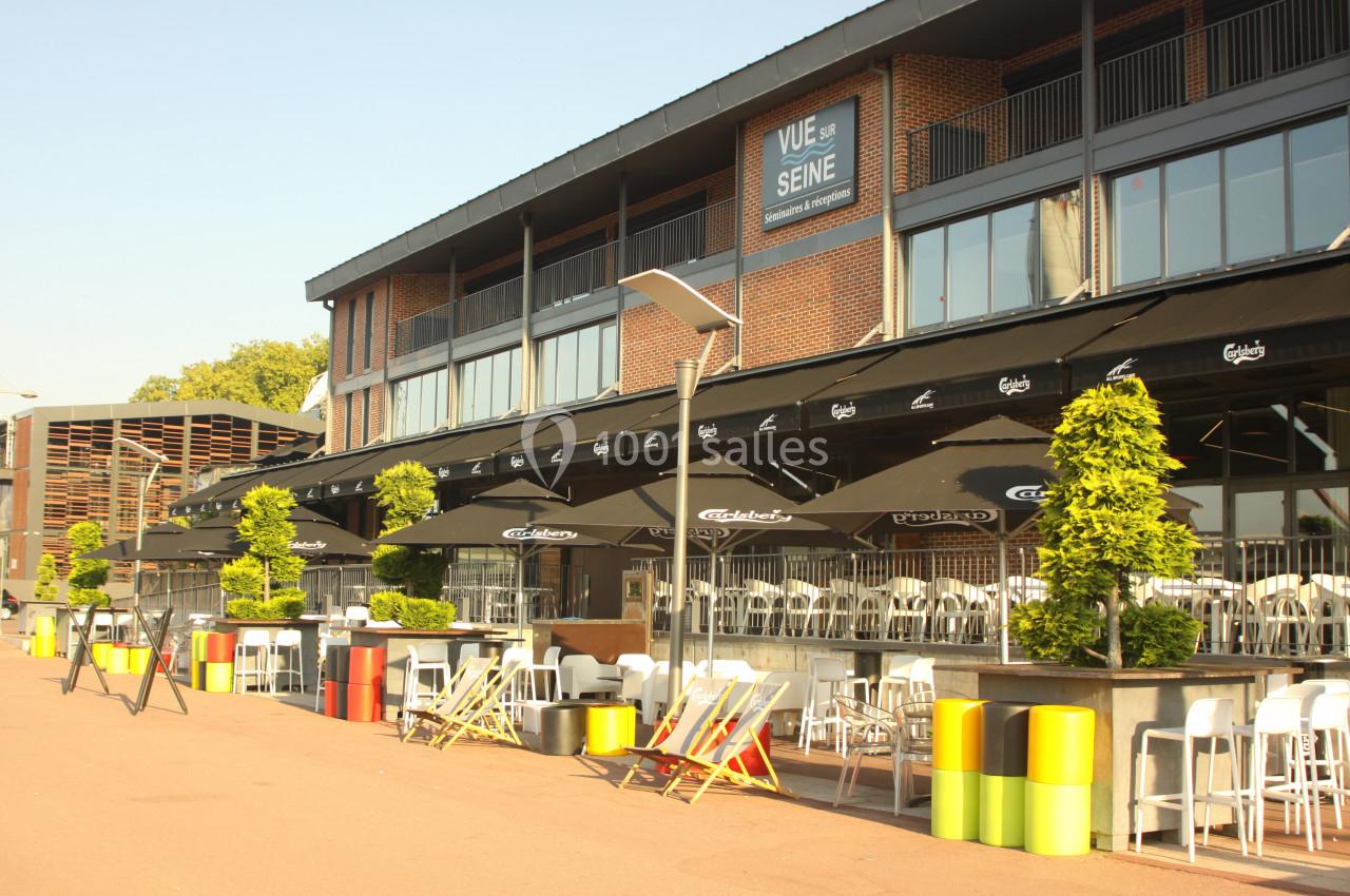 Terrasse ensoleillée avec tables, chaises et parasols devant un bâtiment en briques, bordée de plantes et de mobilier coloré.