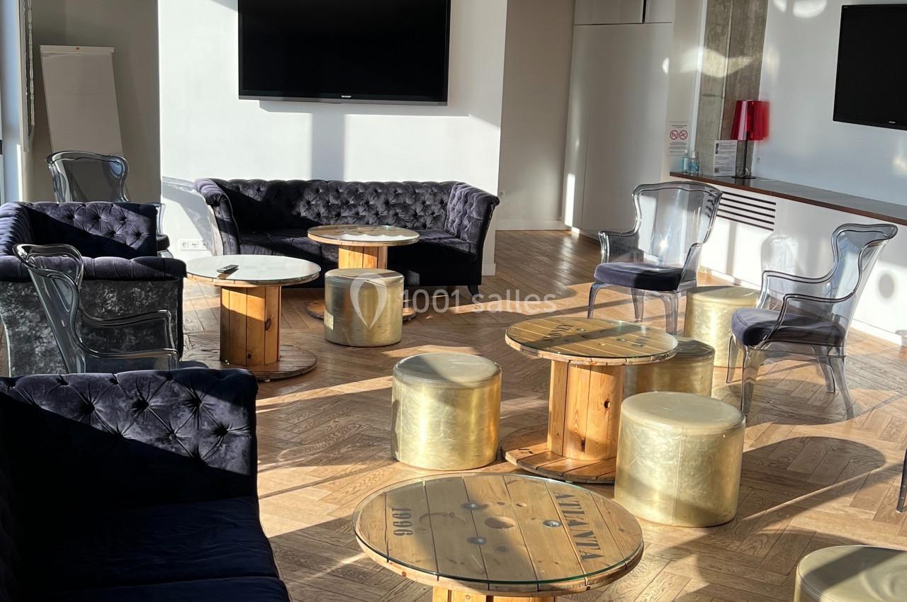 Salle lumineuse avec parquet, canapés en velours, tables en bois et tabourets dorés, éclairée par la lumière naturelle.