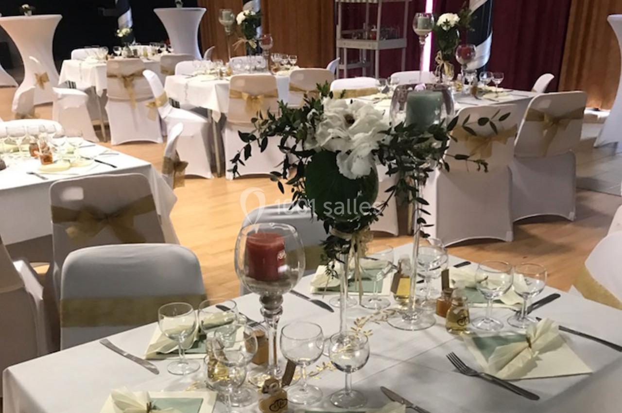 Salle décorée pour un événement, avec tables dressées, nappes blanches, centres de table fleuris et bougies.